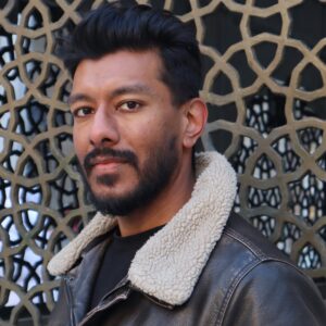Photo of Tanvir Ahmed, a South Asian man with a beard in a bomber jacket. He is standing in front of a thick metal lattice set with the pattern of the khatam sulayman, a popular design motif in Islamic architecture meant to control djinns. © Tanvir Ahmed 2025